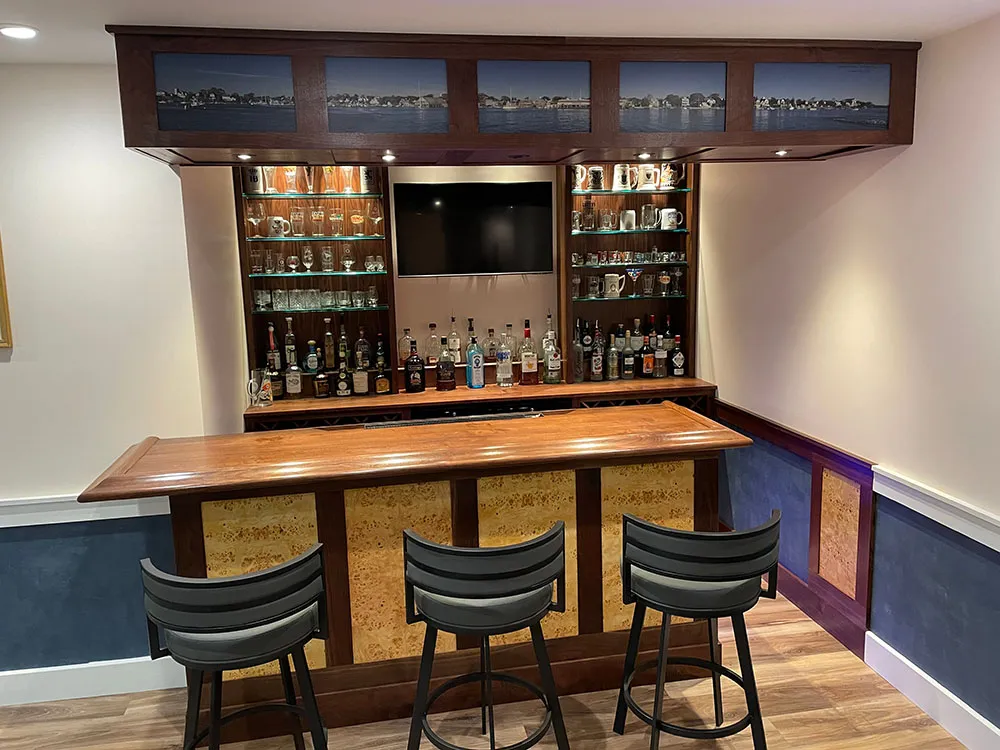 Tan wooden bar with shelves of liquor behind it, and 3 gray bar stools in front, built with materials from Hardwoods Incorporated.